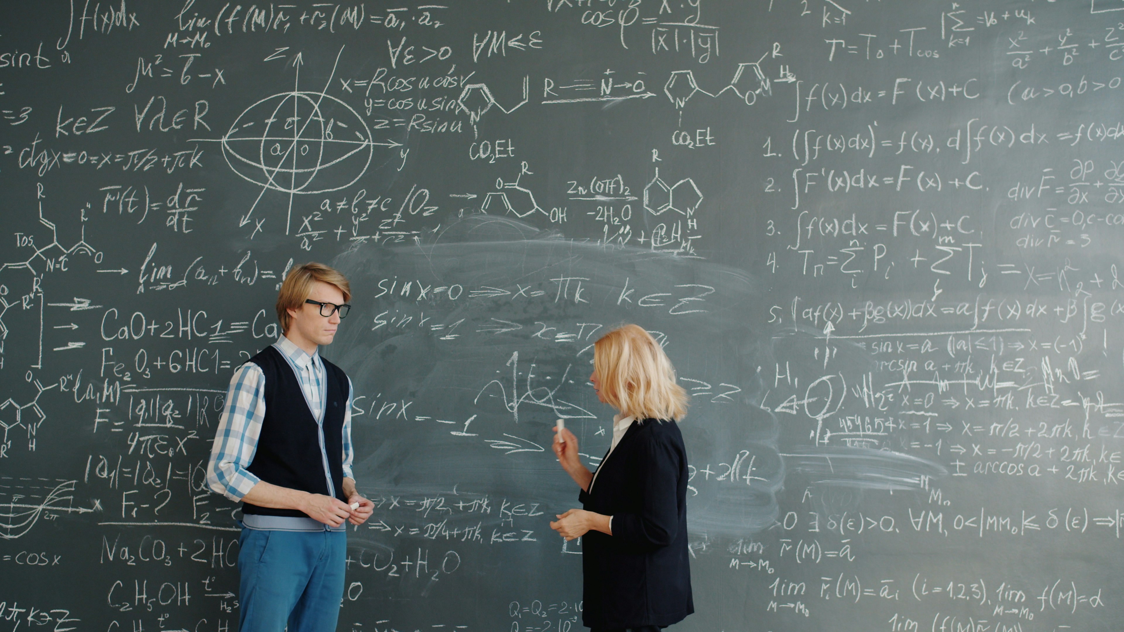 University professor mature lady in suit is teaching handsome guy student writing formulas on chalkboard busy with educational process. People and science concept.